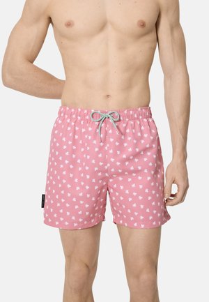Hombre con pantalones cortos de baño rosa con pequeños patrones blancos de pulpos y una cintura ajustable gris, de pie contra un fondo simple.