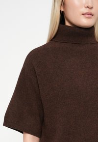 Rudas megztinis su aukštu, susiraukšlėjusiu apykakle, trumpomis rankovėmis ir švelnia, tekstūrine medžiaga. Turi laisvą siluetą ir subtilų, marmurinius raštus.