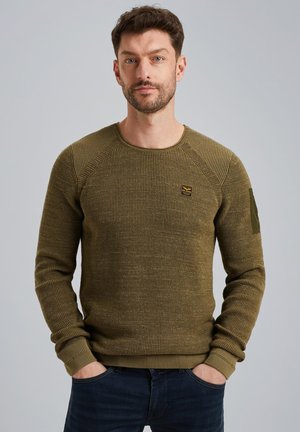 Pull en tricot vert olive avec un col rond, motif texturé et un petit écusson sur la poitrine. Présente des poignets et un ourlet côtelés.