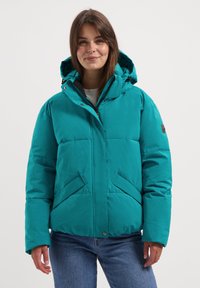 Jonge vrouw in een teal pufferjack met capuchon en blauwe jeans, staand tegen een effen witte achtergrond, met een zachte glimlach.