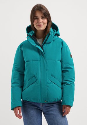 Jonge vrouw in een teal pufferjack met capuchon en blauwe jeans, staand tegen een effen witte achtergrond, met een zachte glimlach.