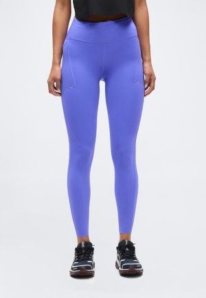 Vrouw die paarskleurige high-waisted legging draagt met zijzakken en zwarte sportschoenen, staand tegen een effen lichte achtergrond.