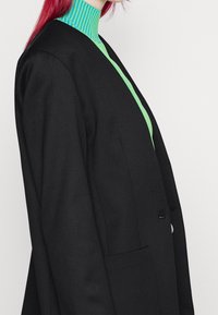 Černý upravený blazer se strukturovaným designem, s klopou a jednou kapsou. Nositý přes svetřík s vysokým límcem pruhovaný v zelené a azurově modré barvě.