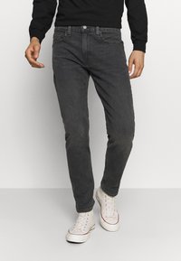 Hombre vistiendo una camiseta de manga larga negra, jeans ajustados gris oscuro y zapatillas blancas de caña alta, de pie contra un fondo liso.