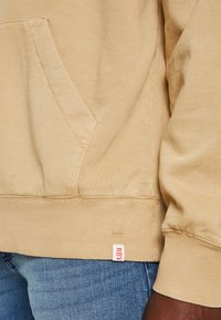 De tan kleurige hoodie heeft een kangoeroe zak, ribgebreide manchetten en een ribgebreide onderkant. Een label met "REV" in het rood is zichtbaar aan de onderkant. Het materiaal lijkt zacht.