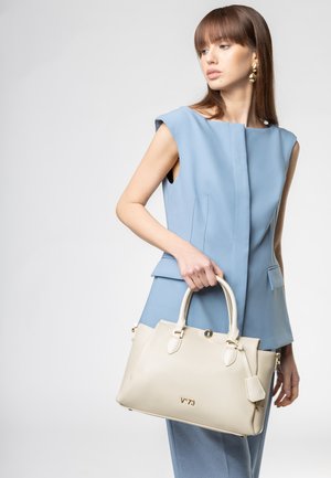 Donna con completo blu senza maniche che tiene una borsa in pelle beige con manici arrotondati e logo dorato "V°73" sul davanti.