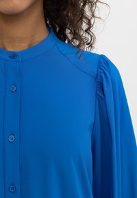 Blusa blu a maniche lunghe con parte anteriore abbottonata, tessuto morbido, spalle plissettate e colletto arrotondato. Texture liscia con vestibilità su misura.