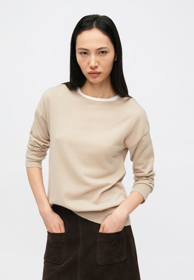 Bēša džemperis ar garām piedurknēm, apaļu kakla izgriezumu un mīkstu tekstūru, kombinēts ar tumši brūnu svārku ar lielām kabatām.