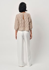 Beige Bluse mit Struktur, Puffärmeln und Schlüsselbeingriff auf der Rückseite, kombiniert mit weißen Weitbein-Hosen. Glattes Material mit dezenten Mustern.