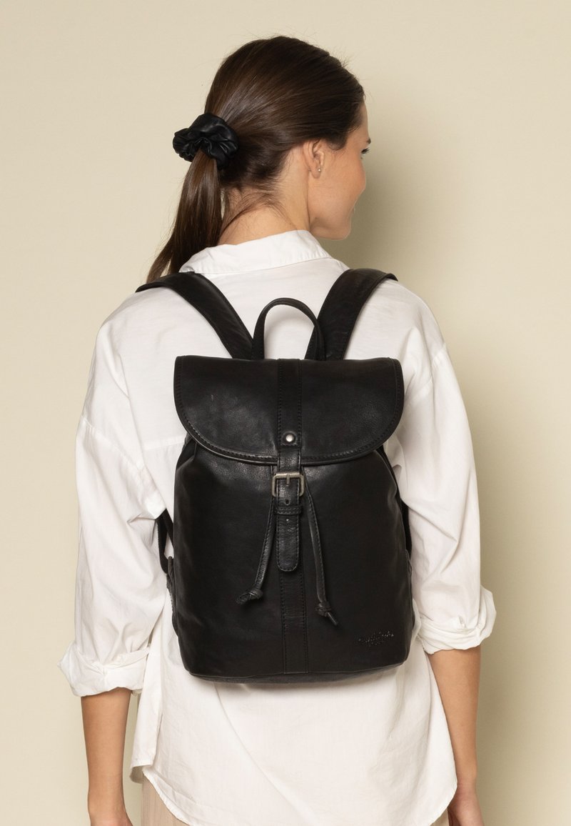 Mochila de cuero negro con solapa redondeada, correas ajustables y cierre de cordón. Textura suave, diseño minimalista y detalles de marca sutiles.