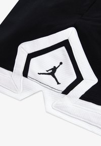 Shorts de sport noirs avec une bordure blanche, présentant un tissu texturé et un logo Jordan noir en position de saut sur un patch blanc.