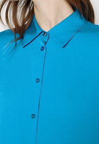 Gros plan sur une personne portant une chemise à col bleu vif avec des boutons visibles et de longs cheveux bruns partiellement visibles.