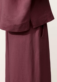 Tenue en lin bordeaux composée d'un haut à manches courtes avec une fente latérale, porté sur une longue jupe avec une poche latérale.