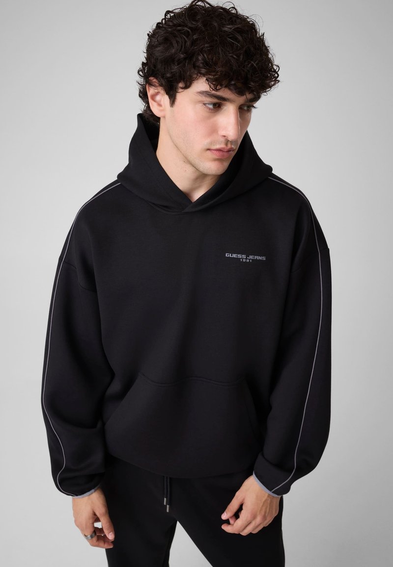 Jeune homme aux cheveux bouclés portant un sweat à capuche noir avec passepoil blanc et le logo "Guess Jeans 1981", regardant vers le bas, debout devant un fond uni.