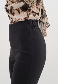 Zwarte, aansluitende broek met een gladde textuur, voorzien van een elastische tailleband en een zijrits, gecombineerd met een transparante top met een patroon.