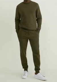 Olivengrünes Sweatshirt- und Jogginganzug-Set aus weichem, strukturiertem Stoff. Mit Rundhalsausschnitt und elastischen Bündchen. Kombiniert mit weißen Sneakers.