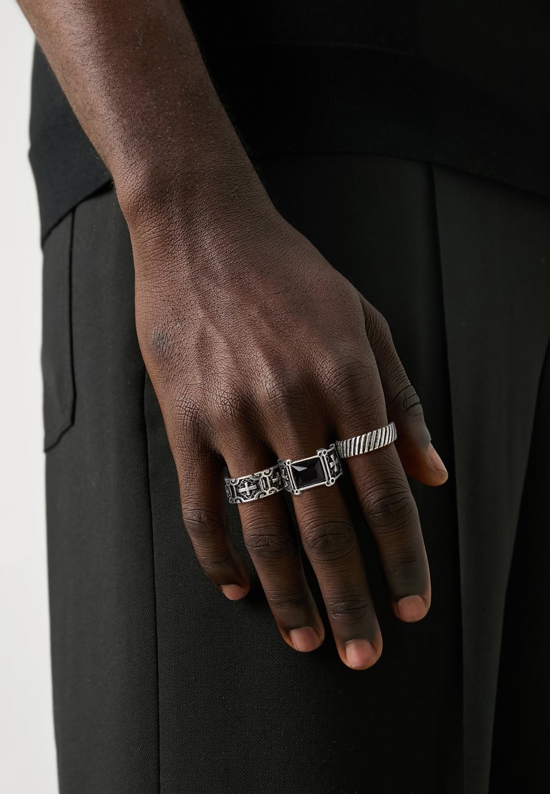 Silver rings on a dark-skinned hand; one features a rectangular black stone, another has intricate patterns, and a third is twisted.