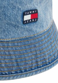 Denim bucket hat in lichtblauw, met gele stiksels en een rechthoekige stofpatch met "TOMMY JEANS" en rood, wit, en blauw accenten.