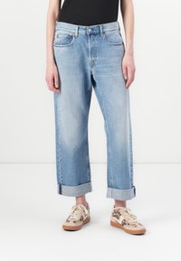 Világoskék, bő szabású farmernadrág, felhajtott szárakkal és öt zsebbel, denim anyagból készült. Mintás bézs és barna sportcipővel viselt.
