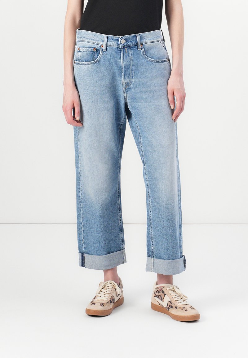 Világoskék, bő szabású farmernadrág, felhajtott szárakkal és öt zsebbel, denim anyagból készült. Mintás bézs és barna sportcipővel viselt.