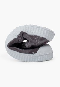 Zapatilla gris de lona sin cordones, plegada para mostrar la suela de goma blanca y la superficie texturizada. Detalles de costura notables y el forro interior son visibles.