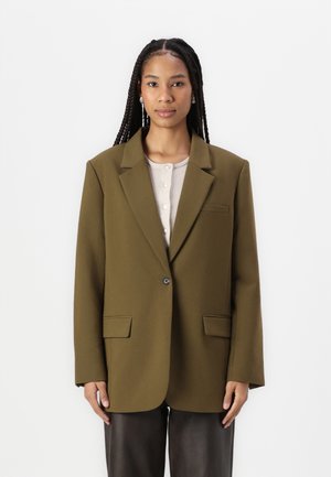 Blazer oversize vert olive avec fermeture à un bouton, revers crantés et deux poches avant, associé à un haut beige clair.