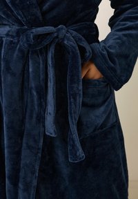 Sotinen tummansininen kylpytakki, jossa on solmittava vyötärö, pitkät hihat ja sivutasku, esitellen pehmeää, samettista pintaa.