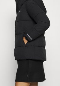 Czarna kurtka puchowa z kapturem, pikowana tekstura, kieszenie na zamek i detal z logo na rękawie. W połączeniu z czarną spódnicą.