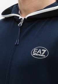 Sudadera con capucha azul marino con cremallera, que presenta un borde blanco en la capucha y el logo "EA7 Emporio Armani". El material tiene una textura suave y un corte ajustado.