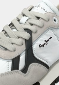 Λευκά και γκρι αθλητικά παπούτσια με υφασμάτινη σουέντ επάνω, λείες δερμάτινες λεπτομέρειες, επίπεδες κορδόνες, μαύρη καουτσούκ σόλα και λεπτομέρεια λογότυπου σε σκούρο καφέ.