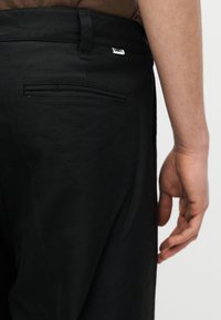 Κοντινό πλάνο ενός ατόμου που φοράει μαύρο παντελόνι, εμφανίζοντας την πίσω τσέπη και το θηλάκι της ζώνης με μια μικρή ορθογώνια ετικέτα μάρκας.