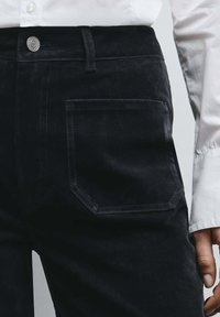 Donkere denim broek met een zichtbare achterzak met een vierkant ontwerp. Materiaal heeft een licht gestructureerd oppervlak. Metalen knoopdetail aan de taille.