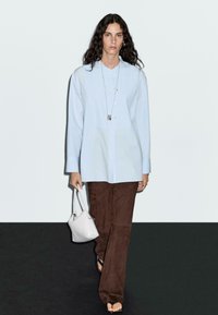 Camicia azzurra chiara con maniche lunghe, abbinata a pantaloni in suede marroni. Una piccola borsa bianca è tenuta nella mano destra.