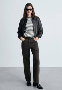 Schwarze Lederjacke über einem gestreiften Langarmhemd, kombiniert mit dunkelbraunen Hosen und schwarzen Schuhen. Das Modell steht mit den Händen in den Taschen.