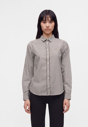 Femme portant une chemise à rayures verticales marron et blanches à manches longues boutonnée, avec un pantalon noir, debout devant un fond blanc uni.