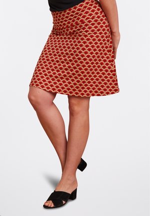 Femme portant une jupe rouge à motifs arrivant aux genoux, un top noir sans manches et des sandales noires à talons carrés et bouts ouverts, debout les jambes croisées.