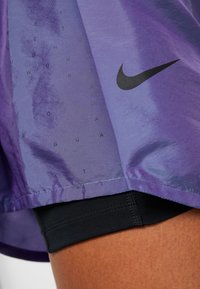Fialové sportovní šortky s lesklou texturou, černým logem Nike a vzorem textu. Černá podšívka viditelná na lemu.