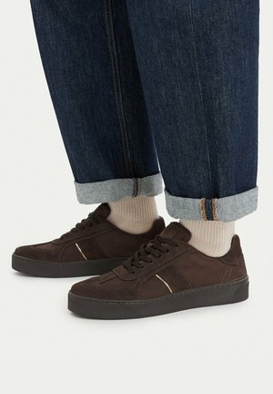 Brune snøresneakers båret med beige sokker og opsmøgede mørkeblå jeans på en neutral baggrund.