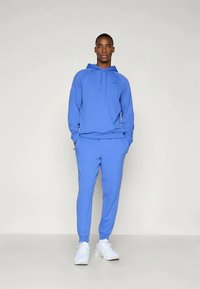 Modèle masculin portant un hoodie bleu et un jogging assorti avec des baskets blanches, se tenant devant un fond clair.