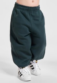 Dunkelgrüne Sweatpants mit elastischem Bund, lockerer Passform und seitlichen Taschen, kombiniert mit weißen Schuhen mit schwarzen Streifen.