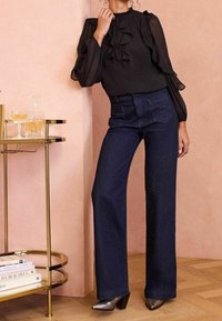 Černá volánková blůza s průhlednými rukávy v kombinaci s tmavě modrými džíny s vysok�ým pasem. Modelka stojí vedle zlatého barového vozíku.