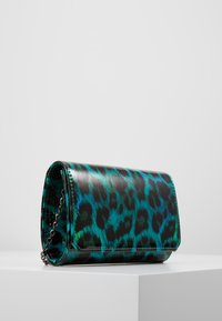 Piccola pochette rettangolare con motivo leopardato turchese e nero e una tracolla in argento, appoggiata su una superficie bianca.