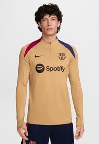 Nike harjoitustoppi vaaleassa sävyssä, jossa on pitkät hihat. Ominaisuuksiin kuuluvat maroonin ja sinisen sävyiset yksityiskohdat olkapäillä, FC Barcelonan logo ja Spotifyn brändäys rintakehässä.