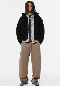 Sort fleece zip-up hoodie over en grønternet skjorte, grå t-shirt, beige sweatpants og mørke sko, med afslappet pasform og casual lag.