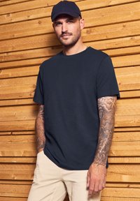 Camiseta de manga corta azul marino, con cuello redondo, fabricada en mezcla de algodón. Presenta un corte relajado y un diseño minimalista. Tatuajes blancos visibles en el brazo.