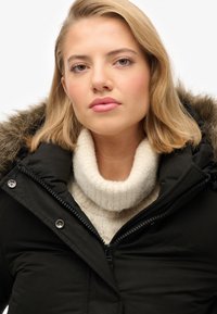 Sort polstret parka med en pelsforet hætte, lynlås foran og stor krave, båret over en kraftig cremefarvet sweater. Glat stoffets tekstur.