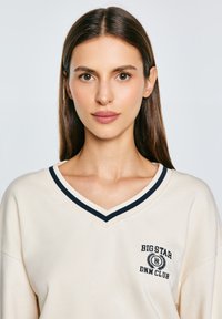 Eine Frau mit langen, glatten braunen Haaren trägt ein cremefarbenes V-Ausschnitt-Shirt mit marineblauen Streifen und dem Text "Big Star DNM Club" auf der linken Brust.
