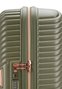 Valigia rigida di colore verde oliva con texture a coste, zip in bronzo, doppio meccanismo di chiusura e etichetta del marchio ben visibile.