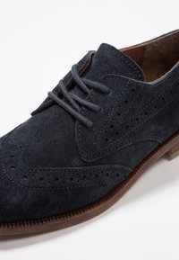 Scarpa in camoscio blu navy con dettagli brogue, chiusura stringata e suola in gomma marrone. Texture liscia e accenti cuciti in tutto.