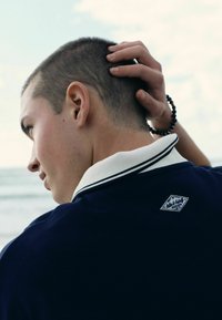 Mladý muž s krátkymi vlasmi, oblečený v tmavomodrej košeli s bielym golierom a korálkovým náramkom, sa dotýka hlavy a stojí pri mori.